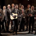 A large band of musicians, holding various instruments, poses together on stage for a group photo.