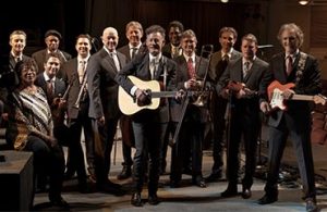 A large band of musicians, holding various instruments, poses together on stage for a group photo.