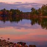 A calm lake reflects autumn trees and a pinkish sky—perfect for leaf peeping near Freeport, Maine.