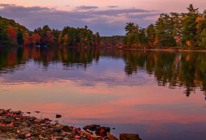 A calm lake reflects autumn trees and a pinkish sky—perfect for leaf peeping near Freeport, Maine.
