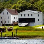 Two white buildings and wooden boats line a grassy shore—classic scenes of vacationing in Maine.