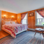 Cozy bedroom with wood-paneled walls, a bed with a red patterned quilt, a rocking chair, and two windows with floral curtains.