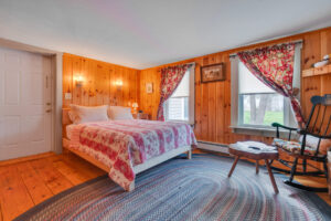 Cozy bedroom with wood-paneled walls, a bed with a red patterned quilt, a rocking chair, and two windows with floral curtains.