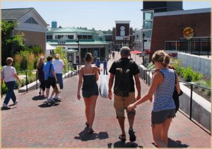 People stroll a sunny shopping area near the clock tower—perfect for itineraries for your James Place Inn vacation in Freeport.